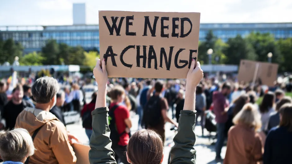 Blick auf eine Protestveranstaltung, viele Menschen von hinten, mittig steht eine Frau und hält ein Plakat mit der Aufschrift "We need a change" hoch